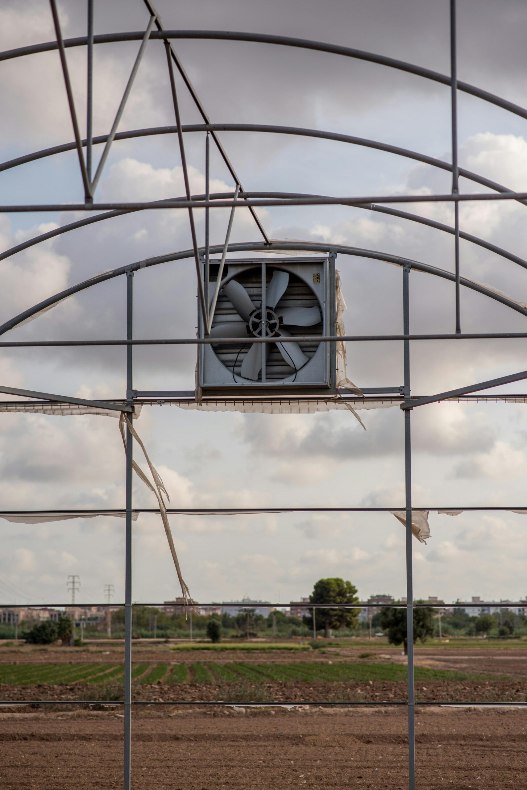 An industrial greenhouse framework with a large ventilation fan, set against a rural landscape.