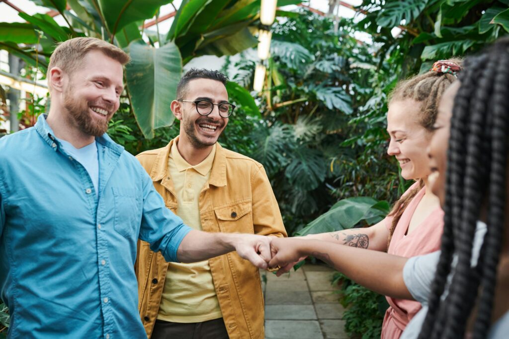 A diverse team of adults enjoying a fist bump gesture in an indoor greenhouse setting.